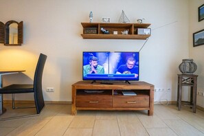 A living room with a wooden TV stand, a flat-screen TV displaying a sports channel, a black chair, and a shelf with decorative items. - Holiday apartment in Büsum with swimming pool (Büsum)