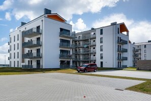 Un edificio residencial moderno de varios pisos con balcones, un camino pavimentado y un automóvil estacionado.