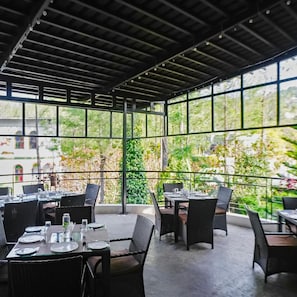An outdoor dining area with tables and chairs, large windows, and a view of greenery.