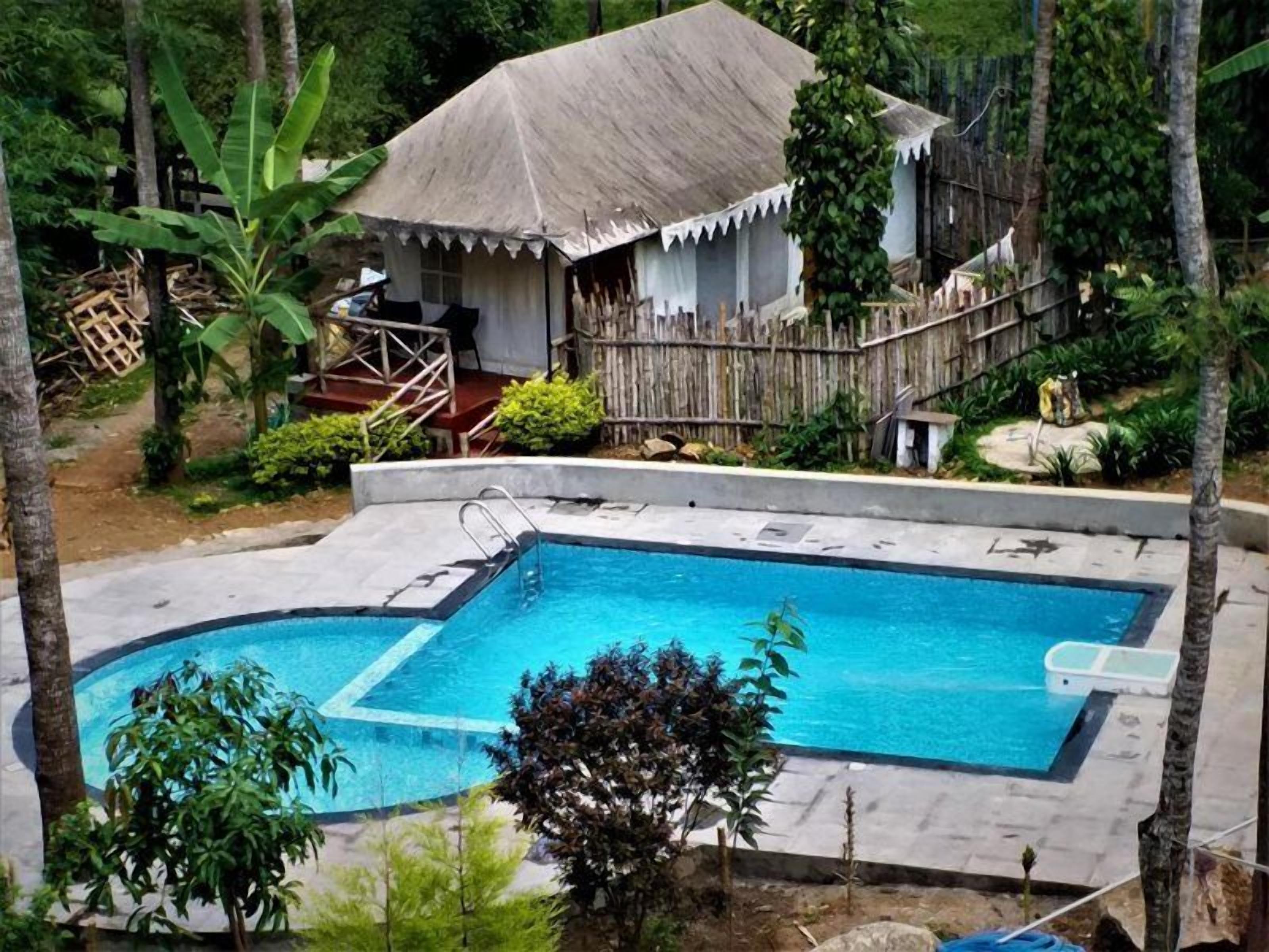 Ein Schwimmbad mit Holzterrasse, umgeben von Bäumen und einem strohgedeckten Gebäude.