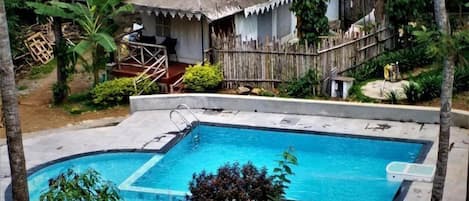 Une piscine dotée d’une terrasse en bois, entourée d’arbres et bordée d’une structure au toit de chaume.