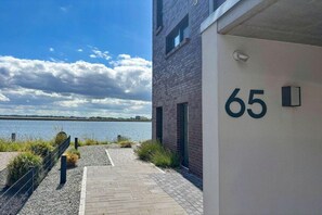 Ein Gebäude mit der Nummer 65, einem gepflasterten Weg und einem Gewässer im Hintergrund.