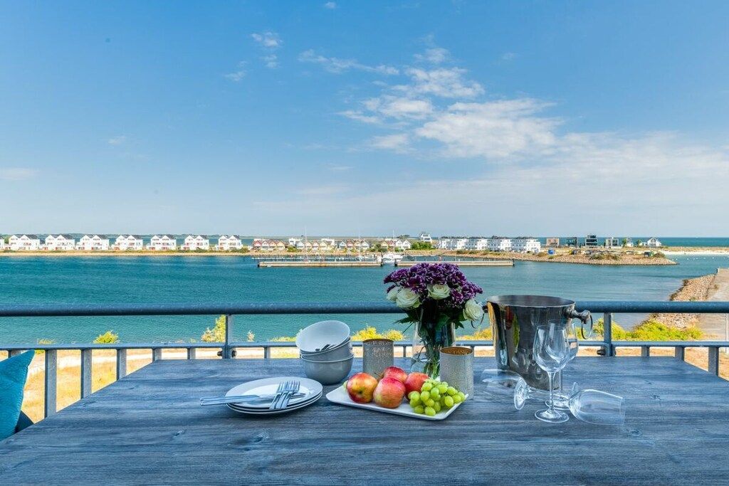 A table with fruits, a vase of flowers, and a wine glass on a balcony overlooking a coastal view.