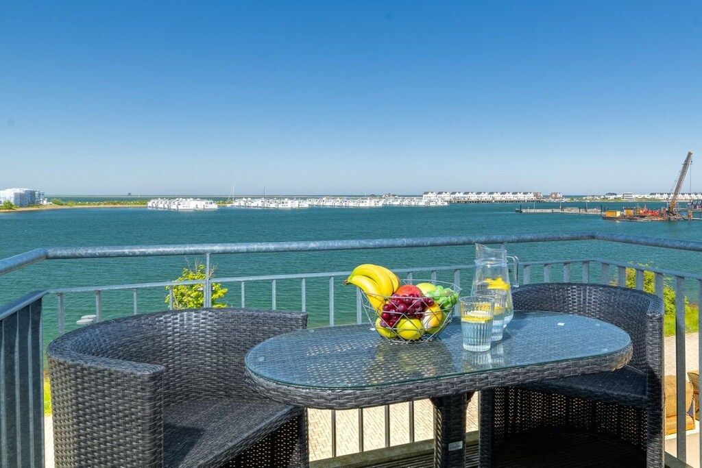 A balcony with wicker furniture and a table with fruit and drinks, overlooking a marina and a pier.