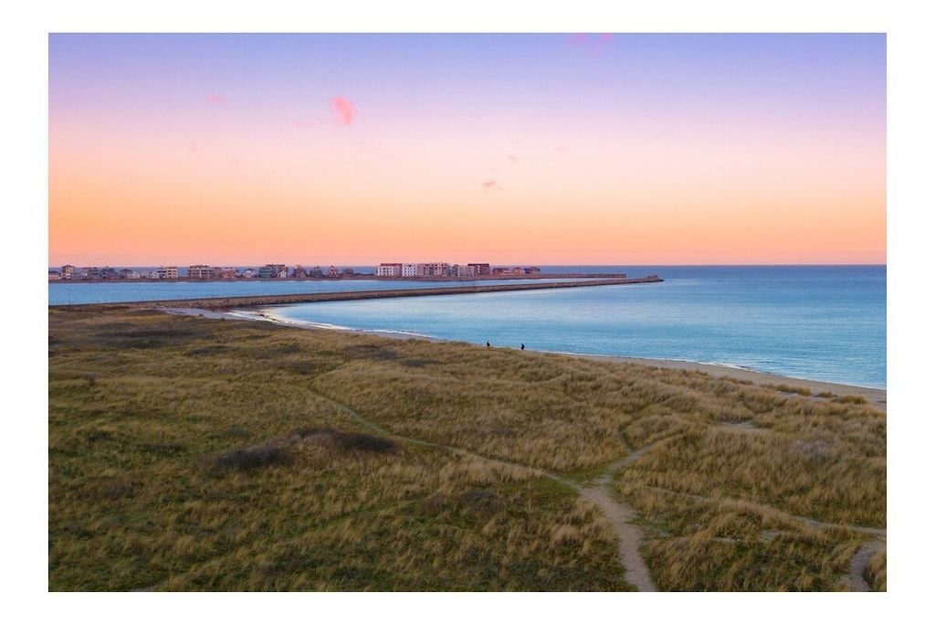A coastal landscape with a clear sky, a sandy beach, and a distant cityscape.