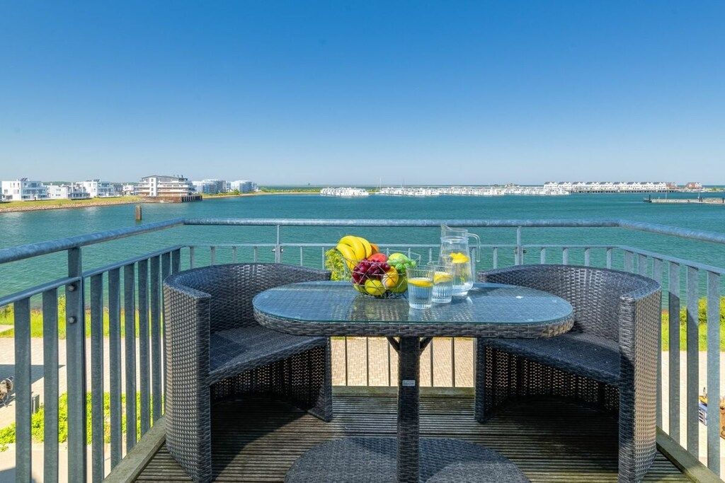 An outdoor dining area with wicker furniture and a table set with fruits and glasses, overlooking a body of water and a distant shoreline.