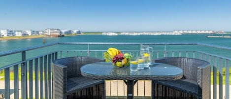 An outdoor dining area with wicker furniture and a table set with fruits and glasses, overlooking a body of water and a distant shoreline.