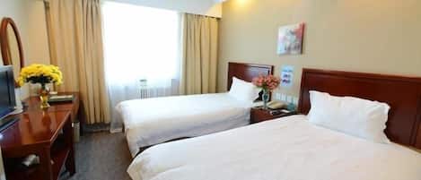 A hotel room with two beds, a desk with a computer, a vase with flowers, and a window with curtains.