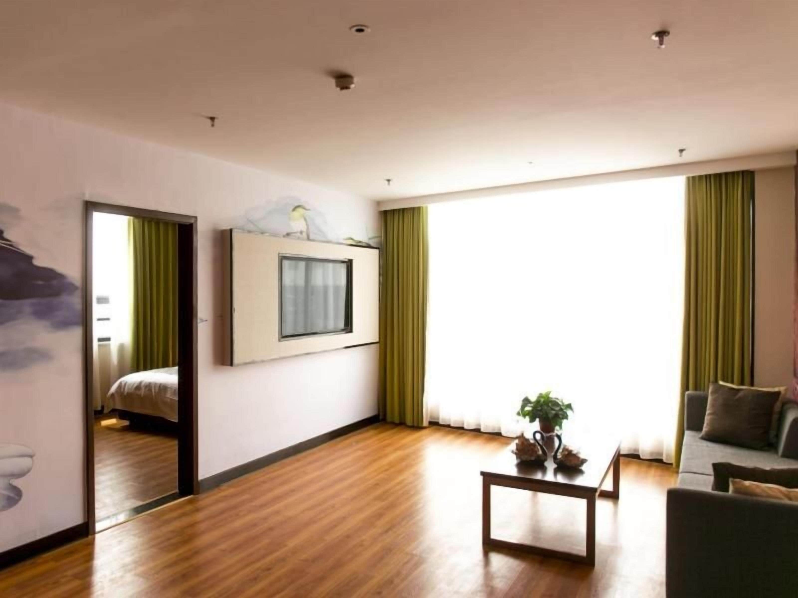 Habitación de hotel con piso de madera, cama con cortina verde, televisor montado en la pared, pequeña mesa con planta y sofá con cojín.