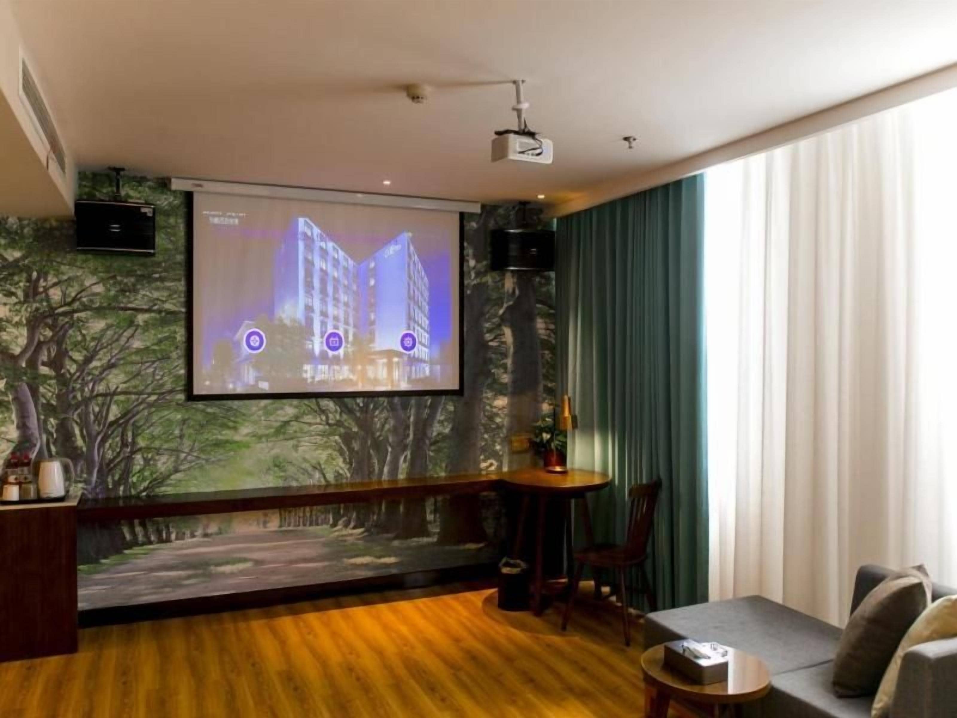 A hotel room with a large screen, a wooden floor, a seating area with a table and chairs, and a view of a building through the window.