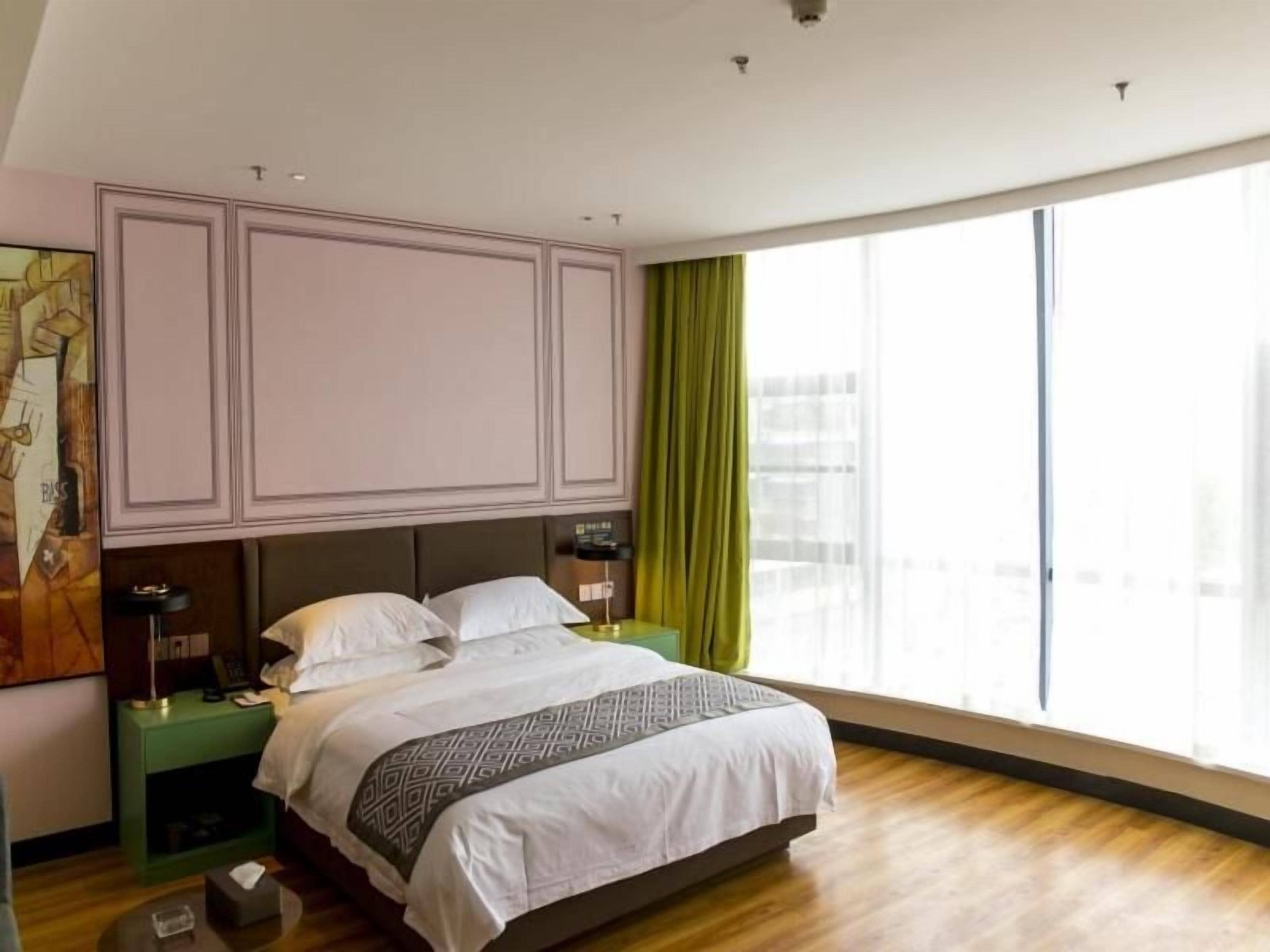Een slaapkamer met een groot bed, een nachtkastje, een lamp en uitzicht op de stad door grote ramen.