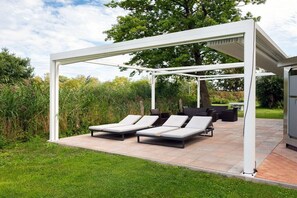 A covered outdoor seating area with lounge chairs and a table, surrounded by greenery. - Holiday home with 1 bedroom (Hohenkirchen Wohlenberger Wiek)