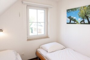 A room with a single bed, a window with a view of a house, and a painting of trees. - Holiday home with 1 bedroom (Hohenkirchen Wohlenberger Wiek)