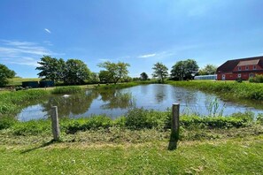 A serene outdoor setting with a pond, greenery, and a red building. - Holiday home with 1 bedroom (Hohenkirchen Wohlenberger Wiek)