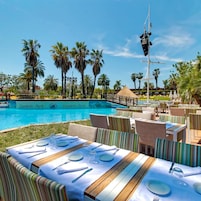 Zona de comedor junto a la piscina con mesas preparadas para dos, con vistas a una piscina con palmeras y cielo despejado.