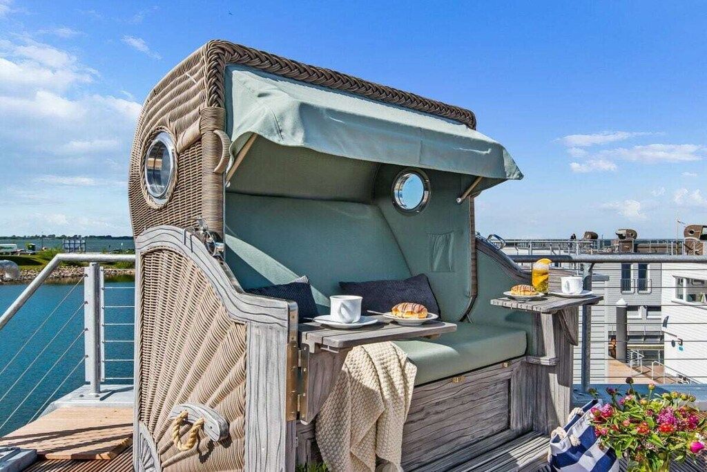 Un espacio exterior acogedor con un banco acolchado, una mesa con una bebida y un pastel, y vista al agua.