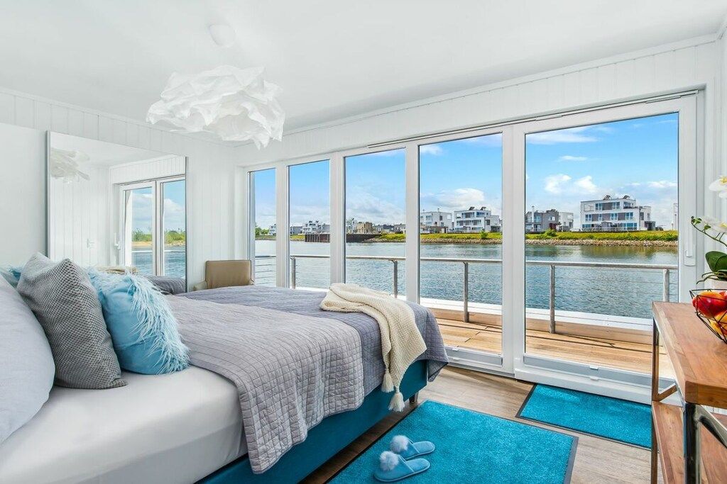 A bedroom with a large window, a bed, a wooden floor, and a view of a body of water and buildings.