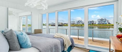 A bedroom with a large window, a bed, a wooden floor, and a view of a body of water and buildings.