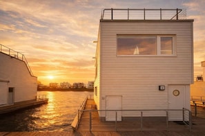 Ein Gebäude am Wasser mit einem Steg, ein Segelboot ist durch ein Fenster sichtbar und im Hintergrund ein Sonnenuntergang.