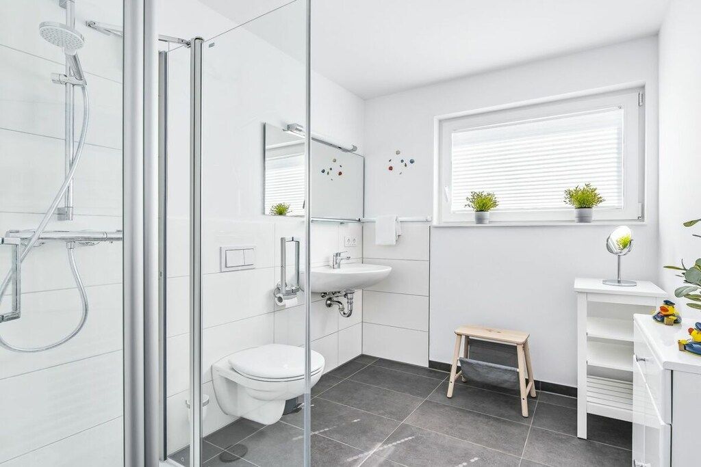 Ein modernes Badezimmer mit Duschkabine aus Glas, einer weißen Toilette, einem Waschbecken und einem kleinen Holzschemel.