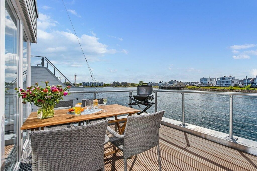 A deck with a table and chairs overlooking a body of water, a grill, and a view of buildings across the water.