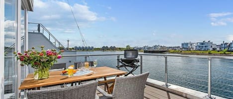 A deck with a table and chairs overlooking a body of water, a grill, and a view of buildings across the water.
