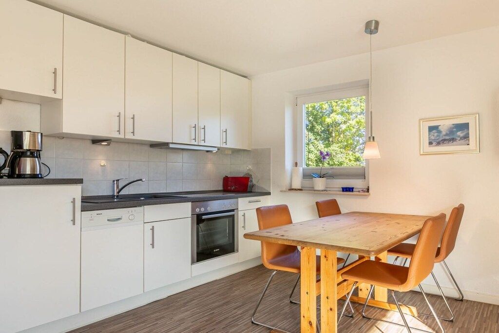 A modern kitchen with white cabinets, a wooden dining table, and a window with a view of greenery.