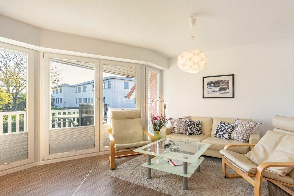 A modern living room with a glass coffee table, beige sofa, and a chair. There is a large window with a view of a building and a tree.
