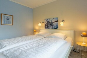 Una cama doble con ropa blanca, dos lámparas de pared y un cuadro con una imagen de un velero.