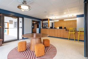 Een moderne hotellobby met een ronde houten tafel, poefs, een receptiebalie en een bar.