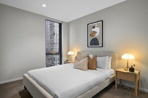 Ein modernes Schlafzimmer mit einem großen Bett, zwei Nachttischen mit Lampen, einem Fenster mit Blick auf ein Gebäude und einem gerahmten Kunstwerk an der Wand.