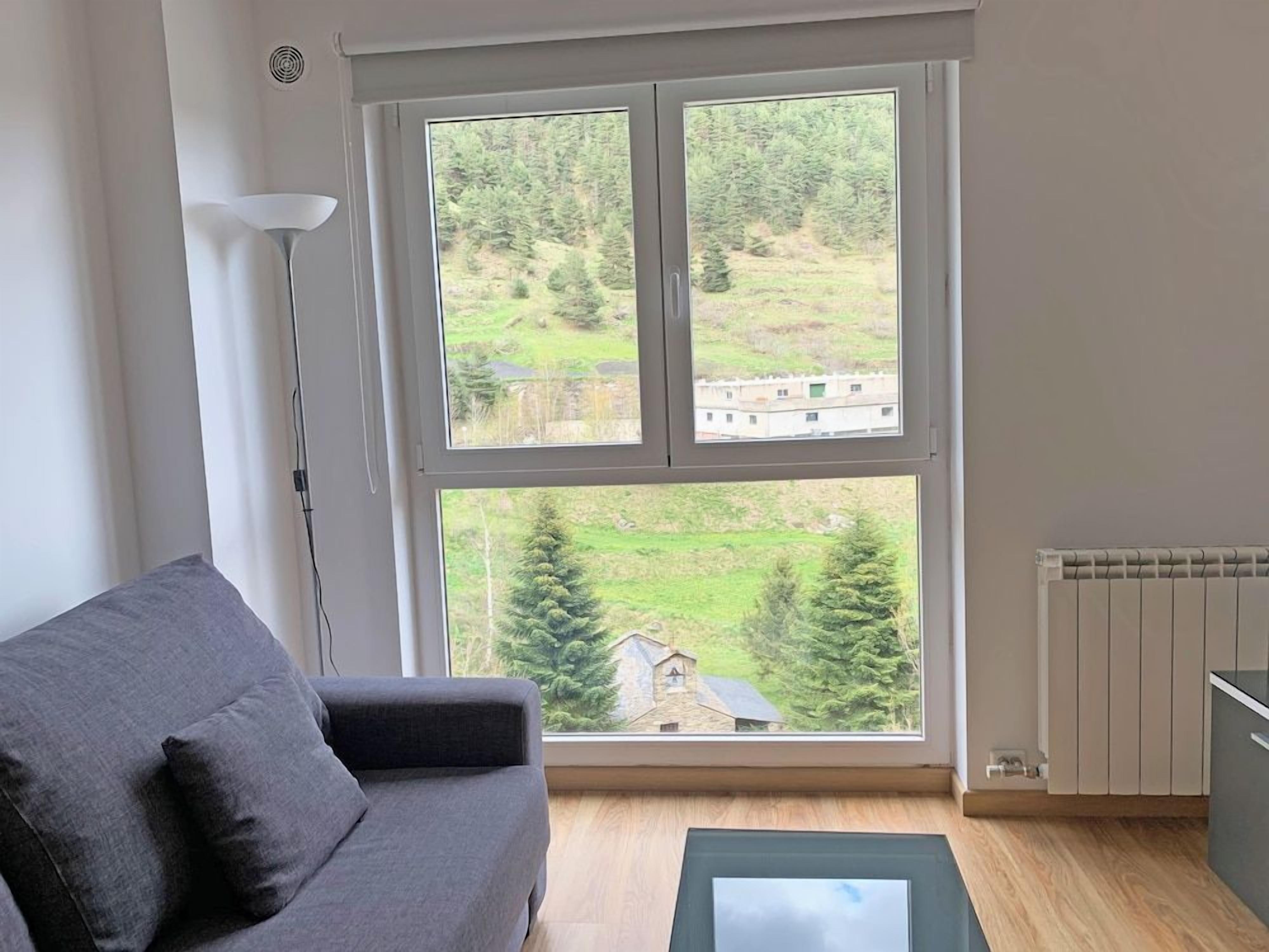 A room with a grey sofa, a floor lamp, and a view of a green landscape through a large window.