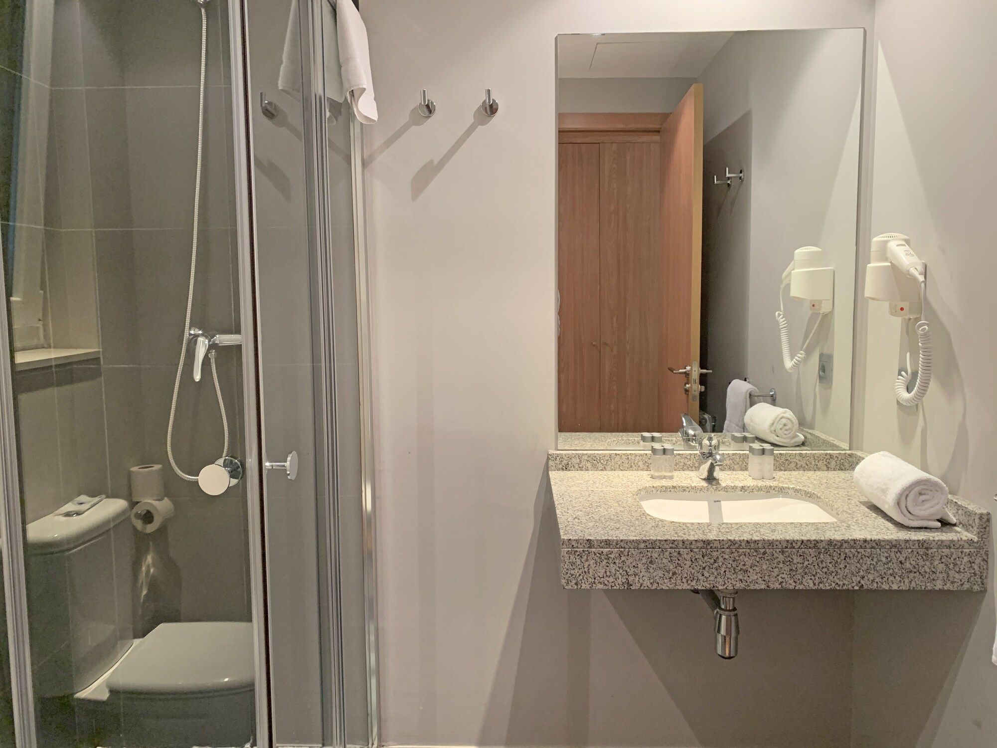 Ein Badezimmer mit Duschkabine aus Glas, Toilette, Waschbecken mit Spiegel und einem an der Wand befestigten Fön.