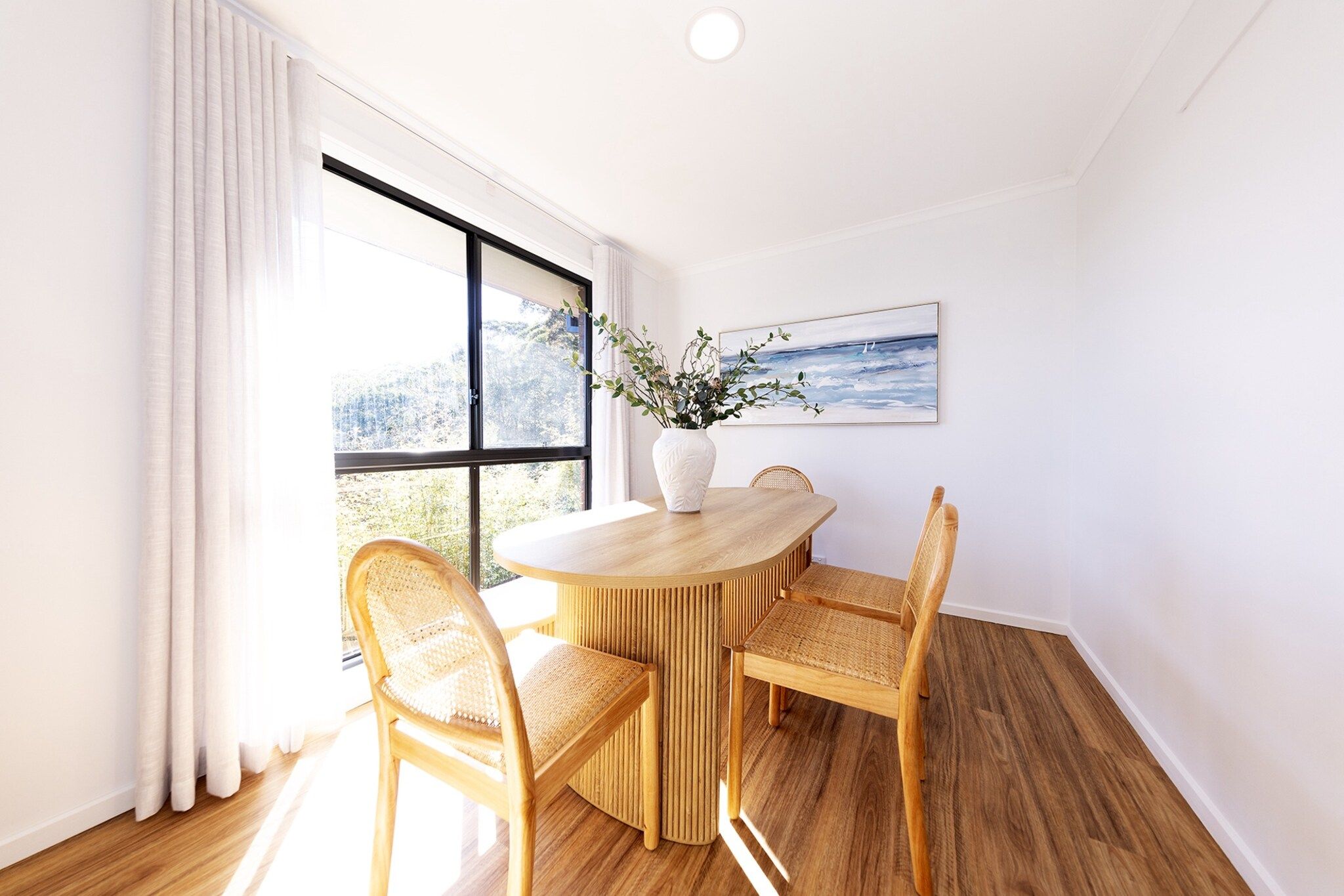 A dining area with a round wooden table, four wicker chairs, a white vase with greenery, and a framed picture on the wall.