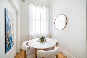 Uma sala de jantar com mesa redonda, quatro cadeiras, espelho redondo e janela com cortinas leves.