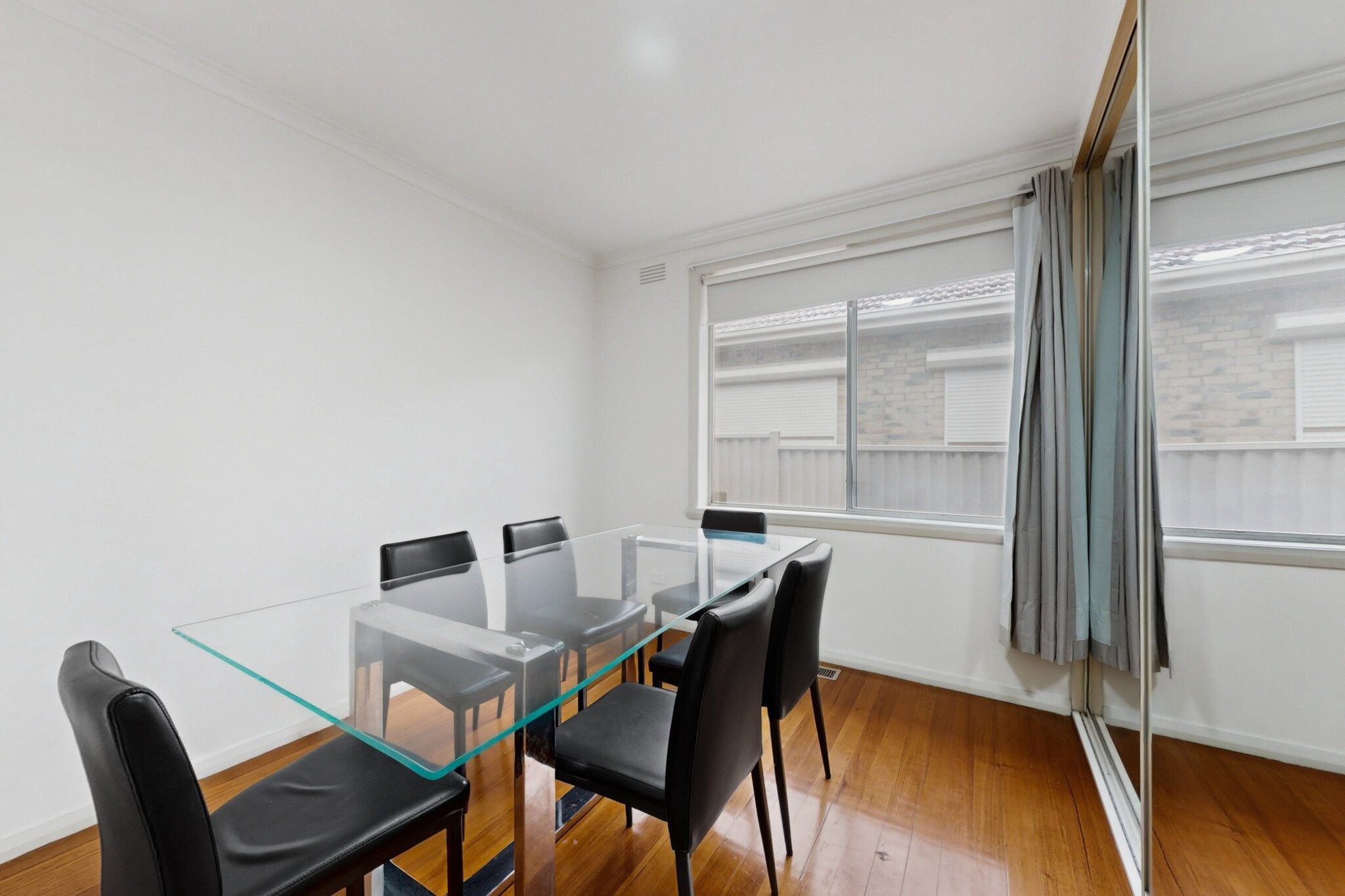 A modern room with a glass-top table and black chairs, large windows with curtains, and a wooden floor.