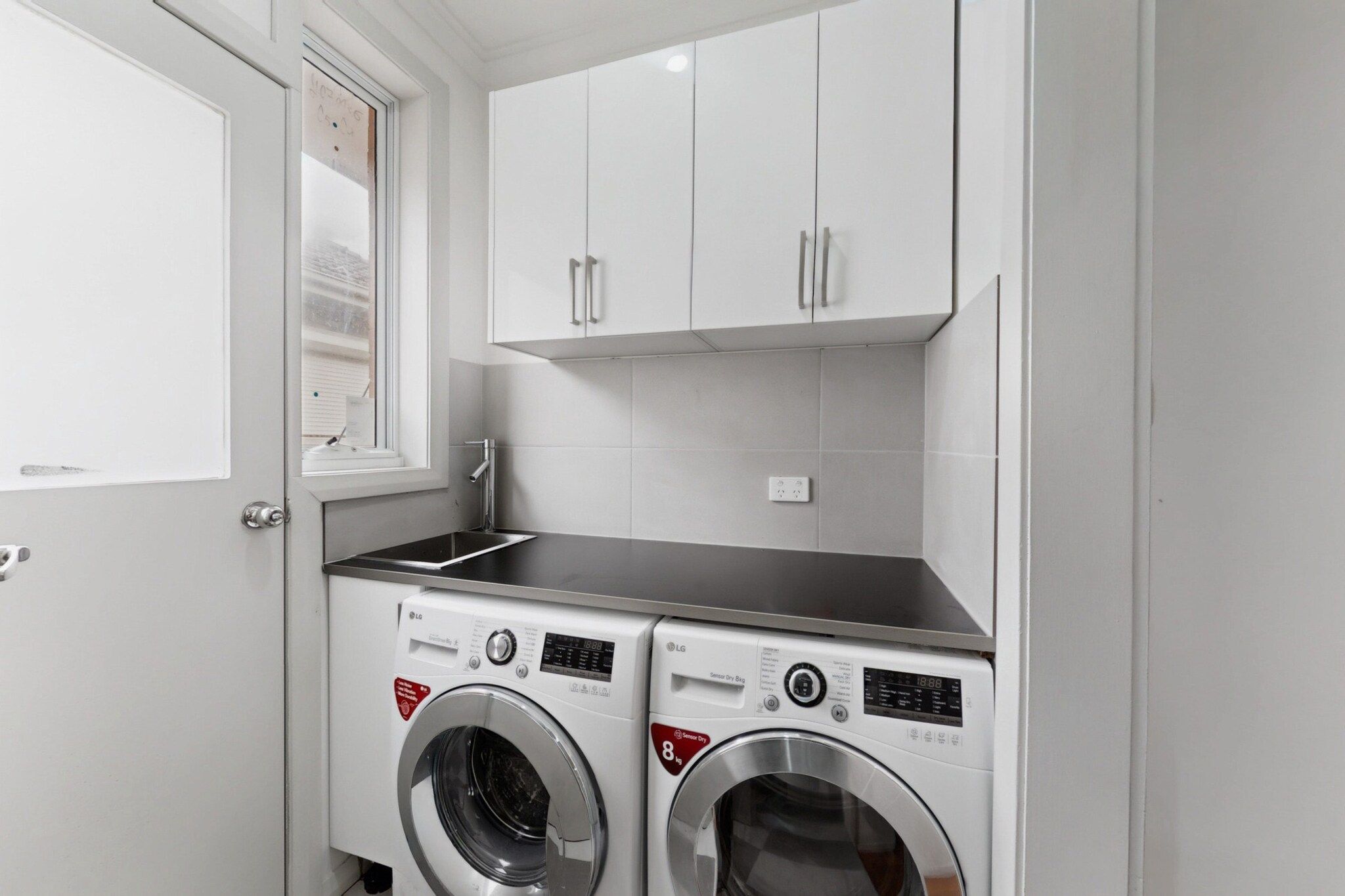 A laundry room with two front-loading washing machines, a window, and white cabinets.