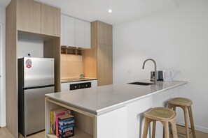 A modern kitchen with a stainless steel refrigerator, a built-in dishwasher, a sink with a faucet, and two wicker stools. - Stylish 1-Bed Apartment With Gym & Pool (Moonee Ponds)