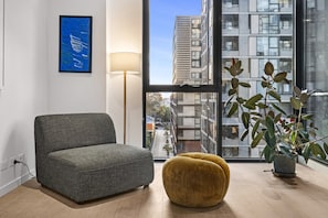 A modern living room with a grey sofa, a yellow pouf, a floor lamp, and a large window with a city view. - Stylish 1-Bed Apartment With Gym & Pool (Moonee Ponds)