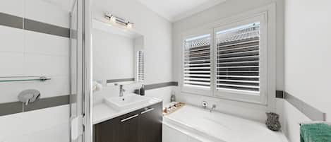 A modern bathroom with a shower, two sinks, a large bathtub, and a window with blinds.