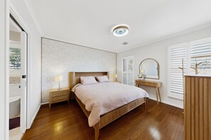 Un dormitorio con cama de madera, lámparas de noche, mesita de noche, cómoda, espejo y ventana con persianas.