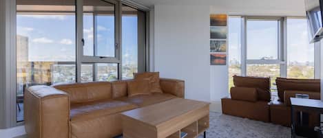 A modern living room with a leather sofa, a wooden coffee table, and a city view through large windows.