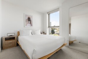 Ein Schlafzimmer mit einem großen Bett, einem Nachttisch, einem Fenster mit Blick auf Gebäude und einem gerahmten Bild.