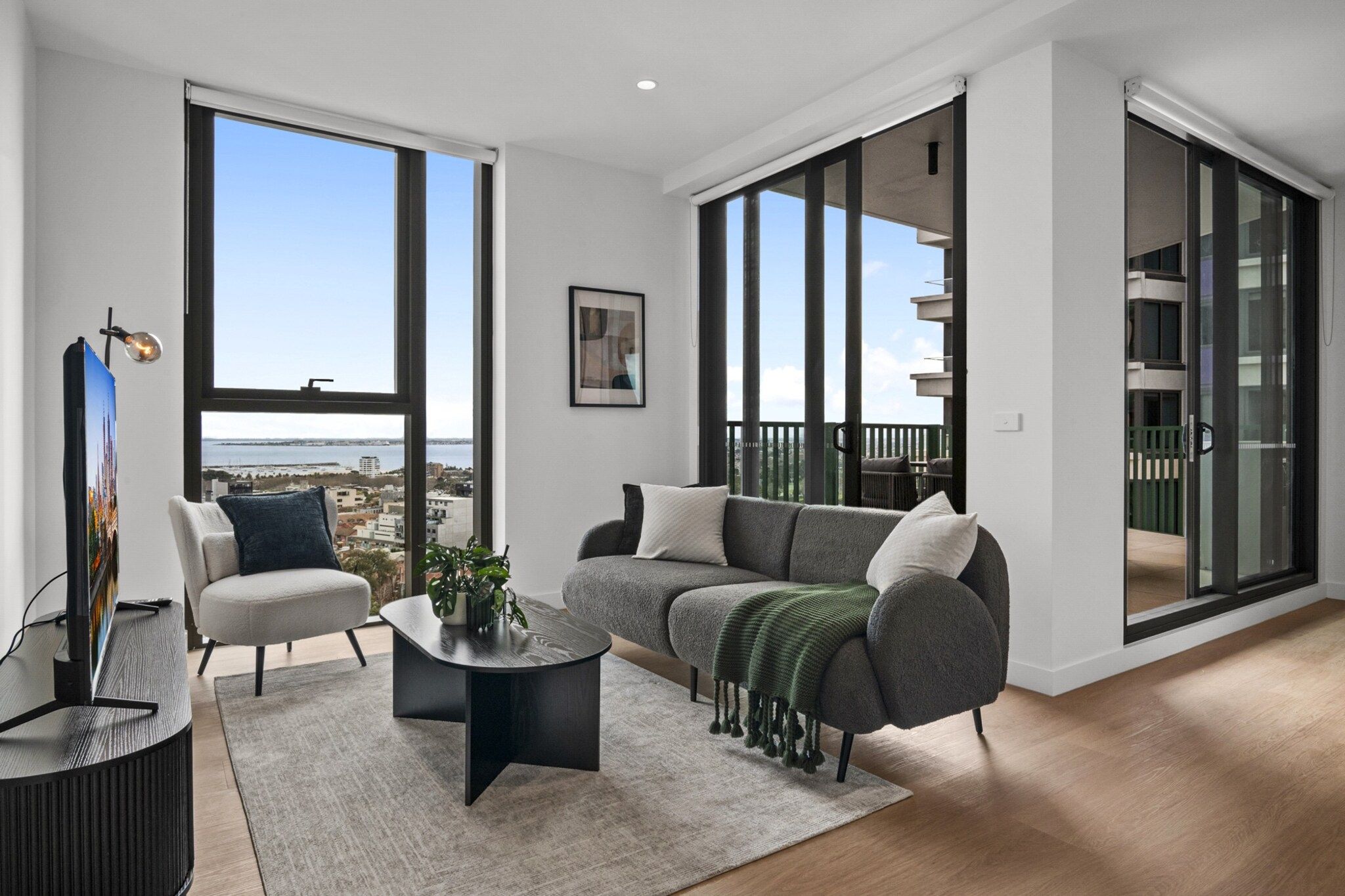 Una sala de estar moderna con sofá, sillón y una mesita de centro, ventanas amplias con vistas a la ciudad y un balcón con barandilla.