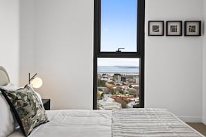 A bedroom with a large window, a bed with a white comforter, a pillow, a bedside lamp, and three framed pictures on the wall. - Light-Filled 3-Bed Brand New Apartment (St Kilda)