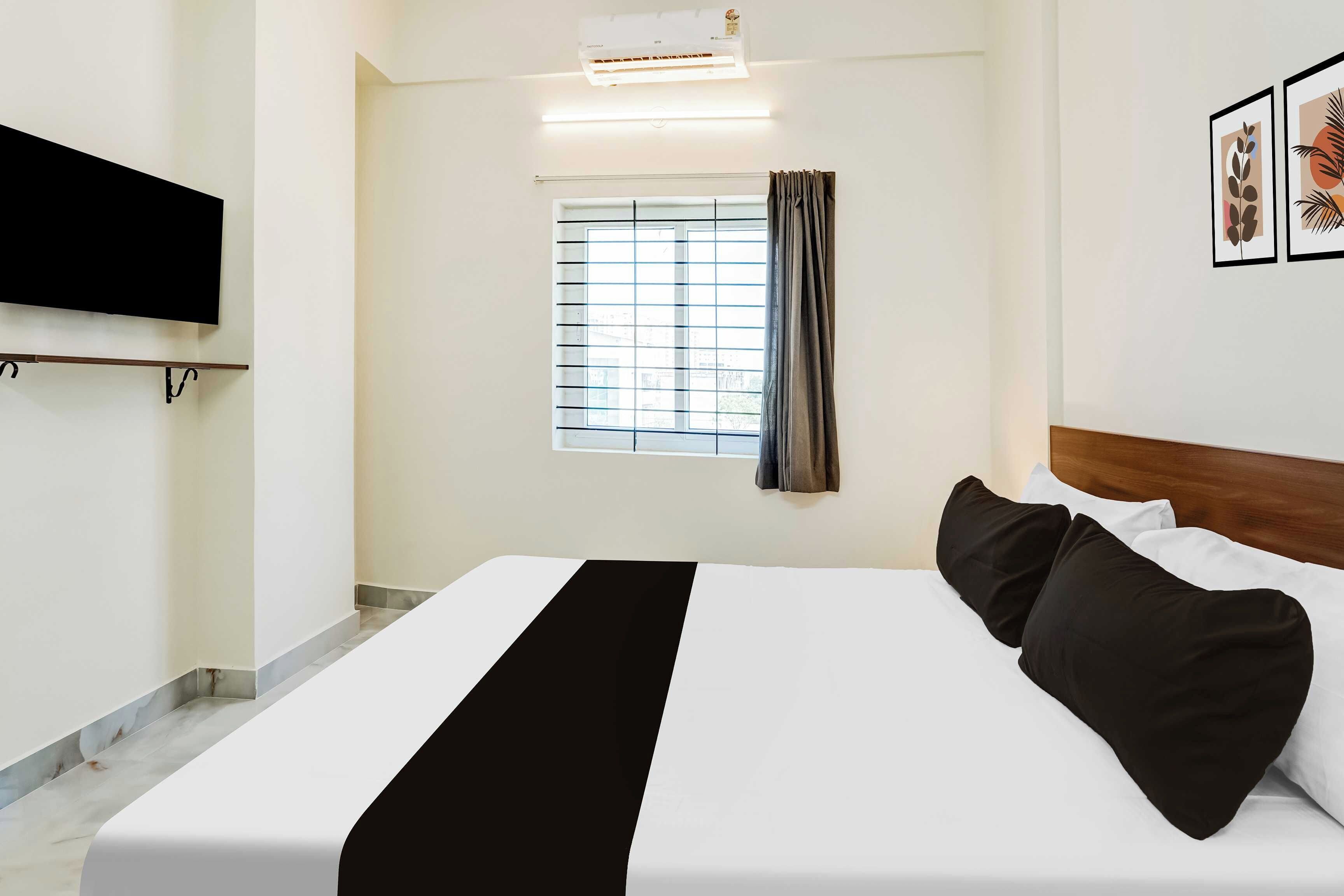 A hotel room with a bed, a flat-screen TV mounted on the wall, a window with curtains, and framed artwork on the wall.