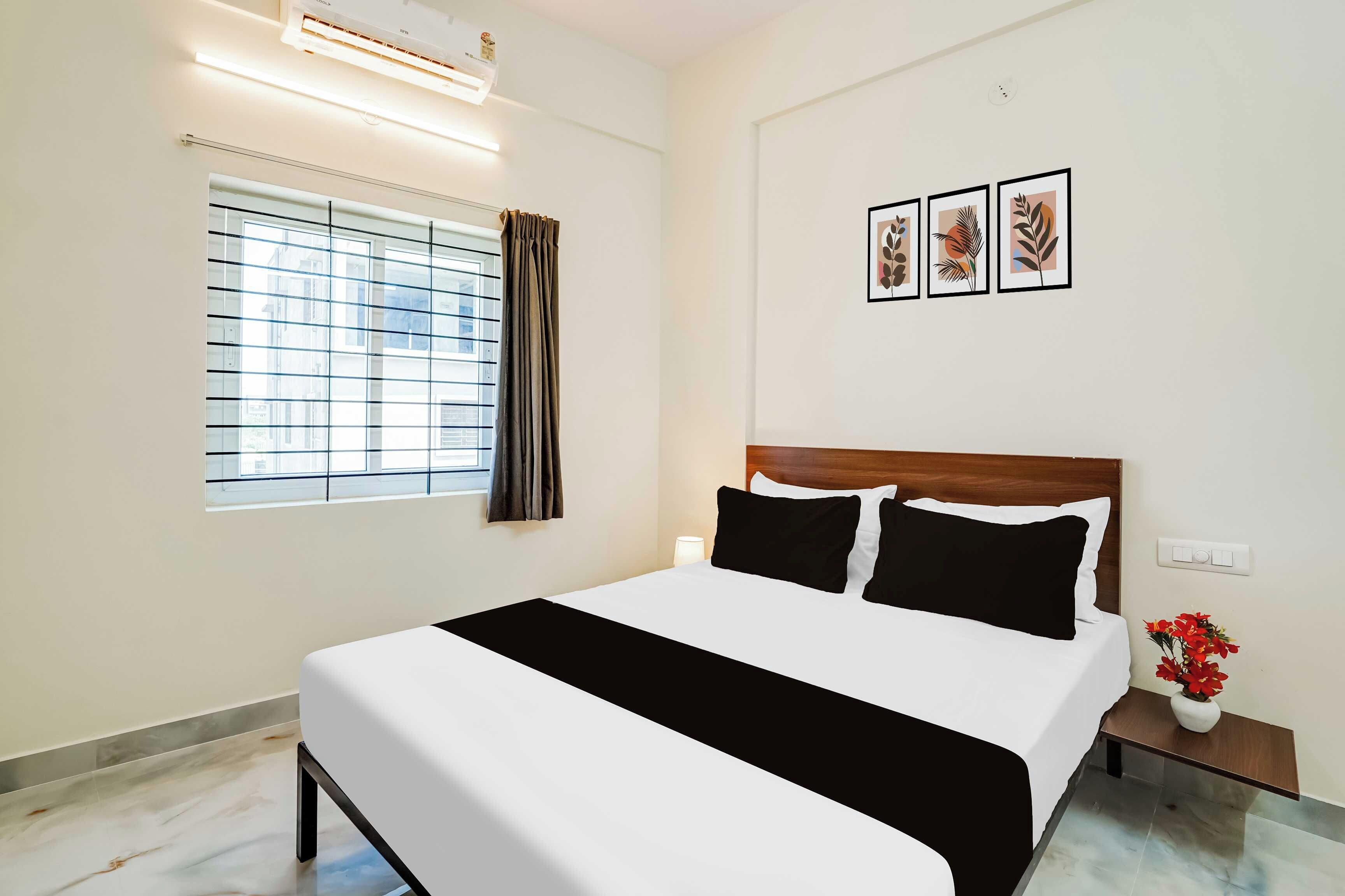 A hotel room with a bed, a window with curtains, a nightstand with a lamp, and a wall with framed artwork.