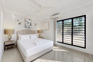 Ein Hotelzimmer mit einem großen Bett, zwei Nachttischen mit Lampen, einem Deckenventilator und einem großen Fenster mit Jalousien.