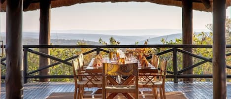 An outdoor dining area with a table set for four, surrounded by a wooden deck and a thatched roof structure.