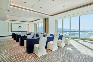 Un salón de conferencias con vista a la ciudad, equipado con mesas, sillas y una pantalla de presentación.